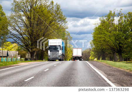 Long haul trucks traveling rural road lined with lush green trees and traditional houses under cloudy spring sky. Heavy freight transport. Long haul trucks traveling rural road lined with lush green trees and traditional houses under cloudy spring sky. Heavy freight transport. 137363298