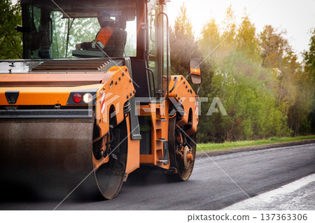 Orange road roller compacting fresh black asphalt during highway construction project. Sunlight flares from trees creating warm light. Orange road roller compacting fresh black asphalt during highway construction project. Sunlight flares from trees creating warm light. 137363306