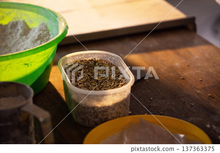Kitchen scene shows preparation of ingredients for baking or cooking process, display of flour in vibrant green bowl, diverse seeds in clear plastic container on rustic wooden table. 137363375