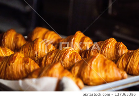 Close up view of numerous freshly baked golden croissants on baking tray, highlighting flaky pastry texture, rich brown crust, fresh from production oven Close up view of numerous freshly baked golden croissants on baking tray, highlighting flaky pastry texture, rich brown crust, fresh from production oven 137363386