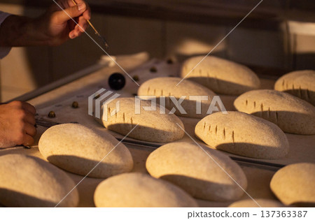 Human hands prepare bread dough loaves for baking process at commercial bakery facility. Manual scoring of dough prior to oven placement. Traditional bread making technique. Human hands prepare bread dough loaves for baking process at commercial bakery facility. Manual scoring of dough prior to oven placement. Traditional bread making technique. 137363387