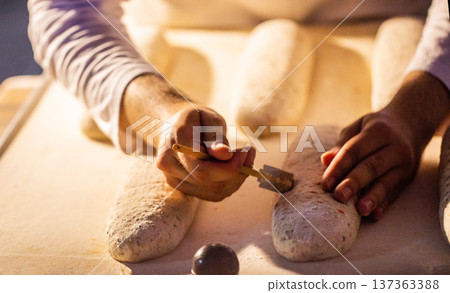Baker hands scoring bread dough loaves with razor tool on floured surface, preparing for baking in bakery or factory, close up view of artisan production of bakery products Baker hands scoring bread dough loaves with razor tool on floured surface, preparing for baking in bakery or factory, close up view of artisan production of bakery products 137363388