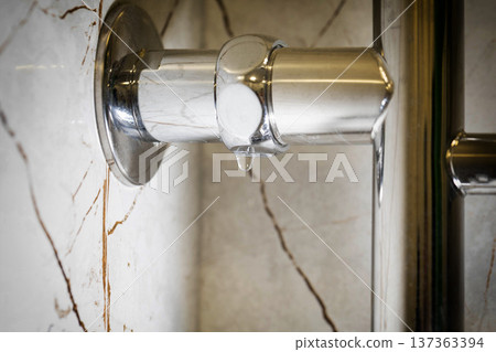 Close up view of water drop leaking from shiny chrome towel warmer pipe in bathroom, showing water damage potential against elegant marble tile wall. Copy space for text Close up view of water drop leaking from shiny chrome towel warmer pipe in bathroom, showing water damage potential against elegant marble tile wall. Copy space for text 137363394