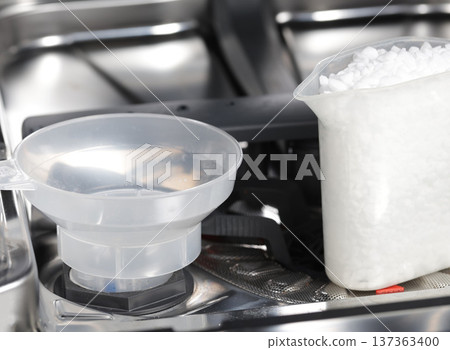 Dishwasher salt refilling procedure inside kitchen appliance, plastic funnel in place, white granules for water softening and machine care Dishwasher salt refilling procedure inside kitchen appliance, plastic funnel in place, white granules for water softening and machine care 137363400