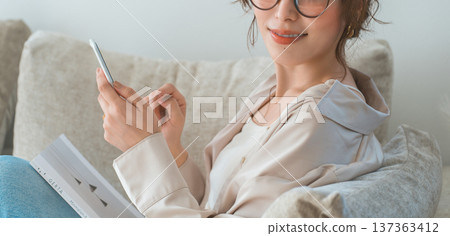 一位亞洲女性坐在客廳的沙發上,看著她的智慧型手機。 一位亞洲女性坐在客廳的沙發上,看著她的智慧型手機。 137363412