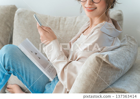 一位亞洲女性坐在客廳的沙發上,看著她的智慧型手機。 一位亞洲女性坐在客廳的沙發上,看著她的智慧型手機。 137363414