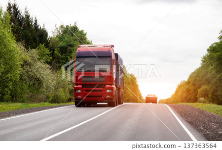 Red semi-truck drives on asphalt highway with white lines, carrying cargo on country road surrounded by green trees and bright sky 137363564