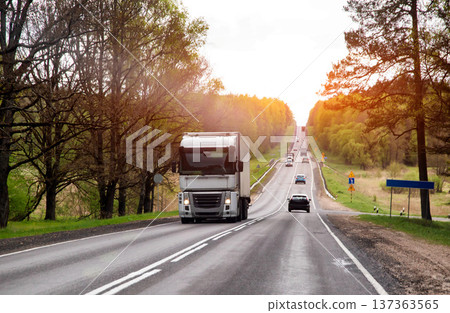Cargo truck and passenger vehicles on two-lane highway, distant golden hour sun illuminating trees and hills, showing road repair and steady traffic flow 137363565