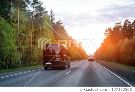 Black cargo van driving on rural road toward horizon light of sunset. Other truck in distance. Lush green forest line road journey freight logistics 137363568