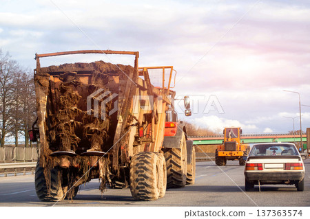 Large agricultural tractor with trailer full of roots and soil travels on multilane highway alongside other vehicles under cloudy sky during daytime. Copy space for text 137363574