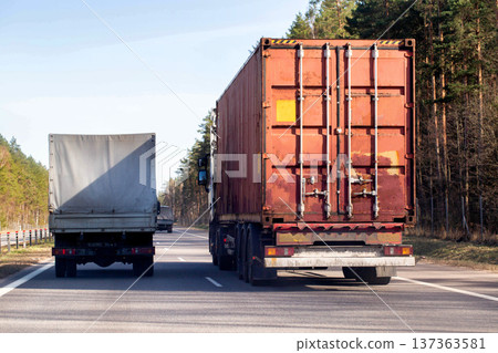 Large semi-trailer truck with red shipping container on highway, smaller truck nearby, commercial transport of goods, logistics, road travel, freight Large semi-trailer truck with red shipping container on highway, smaller truck nearby, commercial transport of goods, logistics, road travel, freight 137363581