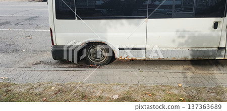 Old white van with flat rear tire parked on urban street sidewalk curb; vehicle body shows extensive rust damage and neglect 137363689
