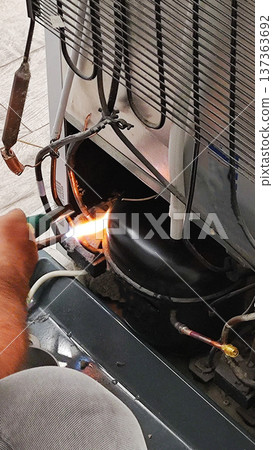 Skillful person hand brazing copper pipe connection of dirty refrigerator compressor unit with intense torch flame during essential repair work indoors Skillful person hand brazing copper pipe connection of dirty refrigerator compressor unit with intense torch flame during essential repair work indoors 137363692