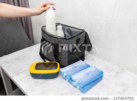 Person hand placing milk bottle into insulated cooler bag with ice packs and lunchbox on table indoors preparing for cold storage keeping food fresh 137363703