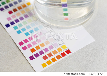 Water quality measurement process using test strip and color comparison chart. Laboratory analysis of water parameters with chemical indicator paper and transparent beaker of liquid. Water quality measurement process using test strip and color comparison chart. Laboratory analysis of water parameters with chemical indicator paper and transparent beaker of liquid. 137363723