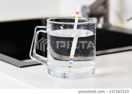 Close up view of test strip immersion in glass mug water showing multiple color sections indicating parameters measurement for water quality analysis on kitchen countertop 137363724