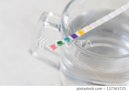 Water quality test strip displaying colorful chemical reaction pads submerged in clear drinking water within transparent glass beaker for parameter analysis and measurement of water purity 137363725