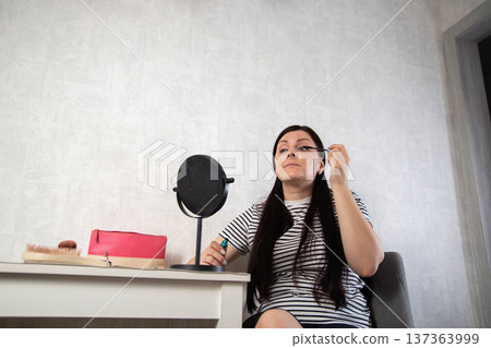 Adult woman carefully applies mascara to upper eyelashes looking into round tabletop mirror, white dressing table with cosmetic bag and brush, indoor beauty routine. Copy space for text Adult woman carefully applies mascara to upper eyelashes looking into round tabletop mirror, white dressing table with cosmetic bag and brush, indoor beauty routine. Copy space for text 137363999