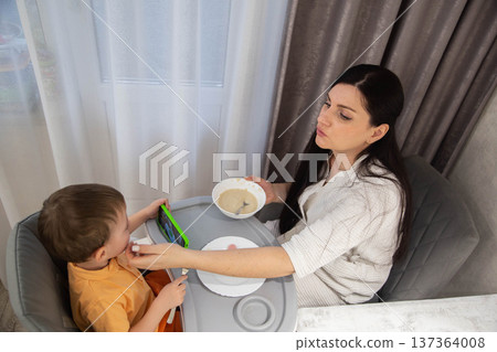 Adult woman feeds young caucasian child in grey high chair, child attention on green smartphone, indoor home mealtime, parenting moment with technology distraction 137364008