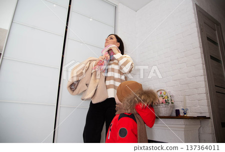 Adult woman in striped sweater, colorful scarf, holding winter coat near closet, young child in red jacket, fur hood, watching woman indoors Adult woman in striped sweater, colorful scarf, holding winter coat near closet, young child in red jacket, fur hood, watching woman indoors 137364011