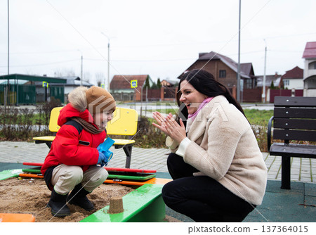 Young boy in red winter coat with adult woman in beige coat laughing and clapping hands at sandbox playtime during overcast day outdoors 137364015