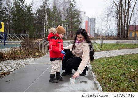 Woman and child sharing tender moment outdoors, boy wearing warm red winter jacket and beanie, woman kneeling near plastic container with colorful objects, parent and kid bonding activity in park Woman and child sharing tender moment outdoors, boy wearing warm red winter jacket and beanie, woman kneeling near plastic container with colorful objects, parent and kid bonding activity in park 137364016