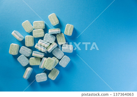 Green and blue dishwasher tablets, individually wrapped, scattered on plain blue background. Cleaning pods for automatic dishwashing machines, top view. Copy space for text 137364063