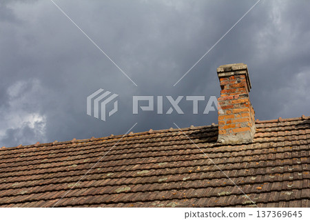 Cloudy sky over a traditional brick chimney on a rustic roof in late afternoon light highlighting the weathered tiles and contrasting textures Cloudy sky over a traditional brick chimney on a rustic roof in late afternoon light highlighting the weathered tiles and contrasting textures 137369645