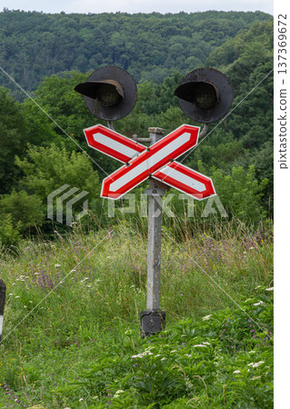 Railroad crossing warning signals stand beside a grassy field with trees in the background on a cloudy day in a rural area Railroad crossing warning signals stand beside a grassy field with trees in the background on a cloudy day in a rural area 137369672