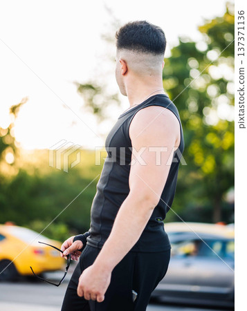 Young man wearing tank top and holding sunglasses on urban street at sunset Young man wearing tank top and holding sunglasses on urban street at sunset 137371136