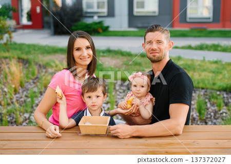 Happy Young Family with two Children Sitting at Wooden Table Outdoors in Summer Happy Young Family with two Children Sitting at Wooden Table Outdoors in Summer 137372027