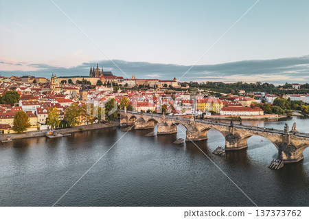 A serene morning scene captures Prague Castle rising above the city, with Charles Bridge stretching over the Vltava River. The old town awakens with soft light and vibrant rooftops. 137373762