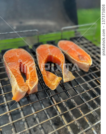 Fresh salmon steaks grilling on a metal grate with smoke and heat, close-up outdoor barbecue scene Fresh salmon steaks grilling on a metal grate with smoke and heat, close-up outdoor barbecue scene 137373808