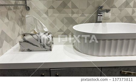 Minimalist bathroom countertop with marble tissue box beside white vessel sink and chrome faucet, geometric tile wall, clean spa-style interior Minimalist bathroom countertop with marble tissue box beside white vessel sink and chrome faucet, geometric tile wall, clean spa-style interior 137373811