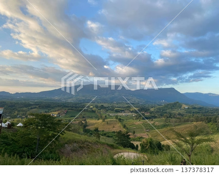 Scenic Mountain Landscape with Rolling Hills and Lush Green Valley under Cloudy Blue Sky Scenic Mountain Landscape with Rolling Hills and Lush Green Valley under Cloudy Blue Sky 137378317