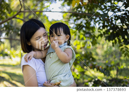 Mother Holding Child Eating Grilled Pork Garden Morning, A warm and authentic photo of a mother cradling her child while enjoying grilled pork outdoors, for food advertising and family documentary. Mother Holding Child Eating Grilled Pork Garden Morning, A warm and authentic photo of a mother cradling her child while enjoying grilled pork outdoors, for food advertising and family documentary. 137378693