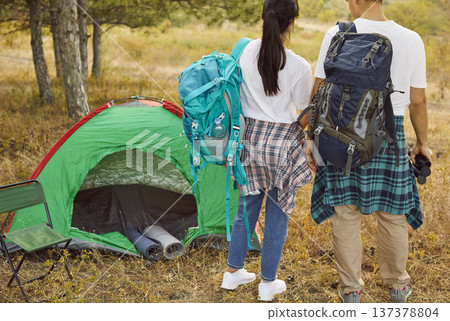 Couple hikers with backpacks by green tent in forest at golden sunset weekend Couple hikers with backpacks by green tent in forest at golden sunset weekend 137378804