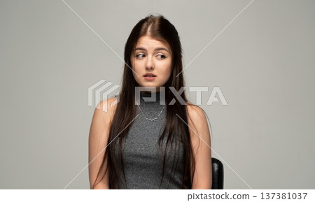 Young woman sitting in studio, expressing deep concern and worry, nervously looking aside with long brown hair 137381037