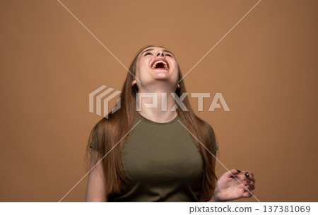 Young woman bursting with laughter, head tilted back, expressing joy and happiness on a brown background Young woman bursting with laughter, head tilted back, expressing joy and happiness on a brown background 137381069