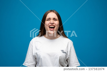 Young woman screaming, expressing anger, frustration, and stress, feeling upset or in pain against a blue studio background Young woman screaming, expressing anger, frustration, and stress, feeling upset or in pain against a blue studio background 137381074
