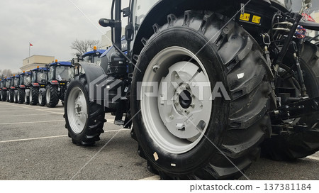 Close up of a large tractor wheel and tire. Row of new blue agricultural tractors parked in a line. Heavy farming machinery industry 137381184