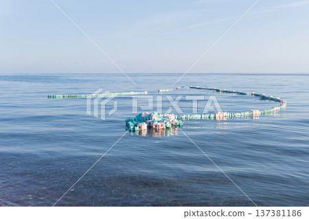 A floating barrier that looks like a snake from a fishing net on the calm blue surface of the sea. Aquaculture equipment with foam buoys in ocean water 137381186
