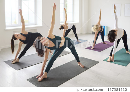 Diverse group of women practicing triangle yoga pose in bright modern studio Diverse group of women practicing triangle yoga pose in bright modern studio 137381310