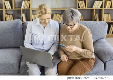 Senior woman paying online with help, credit card and laptop on sofa at home Senior woman paying online with help, credit card and laptop on sofa at home 137382380