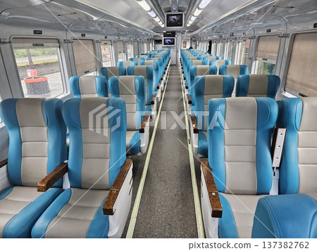 Interior view of a modern clean train carriage with comfortable blue and grey ergonomic seating, featuring bright fluorescent lighting, overhead monitors, and large windows showing green scenery. Interior view of a modern clean train carriage with comfortable blue and grey ergonomic seating, featuring bright fluorescent lighting, overhead monitors, and large windows showing green scenery. 137382762