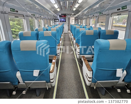 Interior view of a modern clean train carriage with comfortable blue and grey ergonomic seating, featuring bright fluorescent lighting, overhead monitors, and large windows showing green scenery. Interior view of a modern clean train carriage with comfortable blue and grey ergonomic seating, featuring bright fluorescent lighting, overhead monitors, and large windows showing green scenery. 137382770