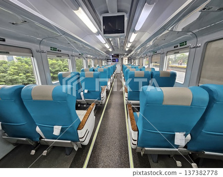 Interior view of a modern clean train carriage with comfortable blue and grey ergonomic seating, featuring bright fluorescent lighting, overhead monitors, and large windows showing green scenery. Interior view of a modern clean train carriage with comfortable blue and grey ergonomic seating, featuring bright fluorescent lighting, overhead monitors, and large windows showing green scenery. 137382778