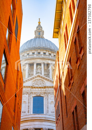 Visitors walk along a narrow street in London, looking up at St Pauls Cathedral. The sunlight highlights the dome and architectural details of the building. 137382798