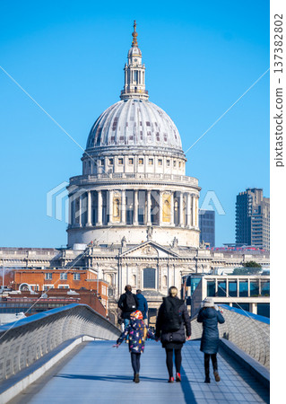 People walk across Millennium Bridge towards St Pauls Cathedral in London. The dome of the cathedral stands prominently against a clear blue sky. 137382802