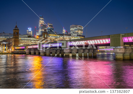 Cannon Street Railway Bridge stands over the water at night. The bridge is lit with colorful lights. The skyline of London features tall buildings in the background. Cannon Street Railway Bridge stands over the water at night. The bridge is lit with colorful lights. The skyline of London features tall buildings in the background. 137382807